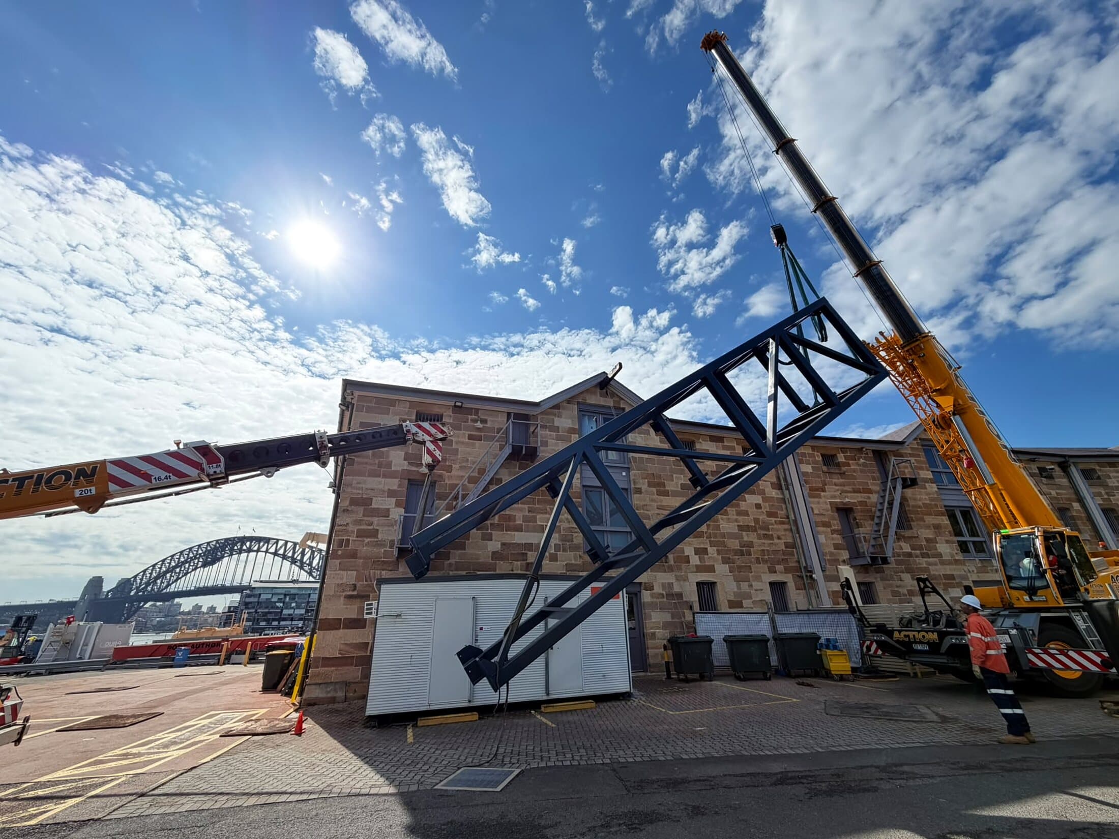 Fabricated steel lift shaft being lifted by crane beside a sandstone building, with the Harbour Bridge behind